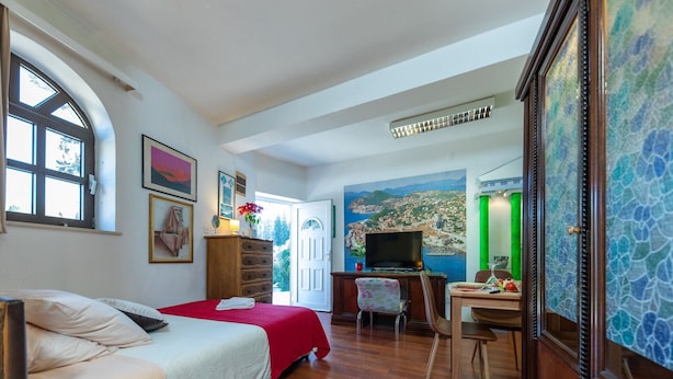 A bedroom with a bed, a desk, a television, and a mural of a coastal town.