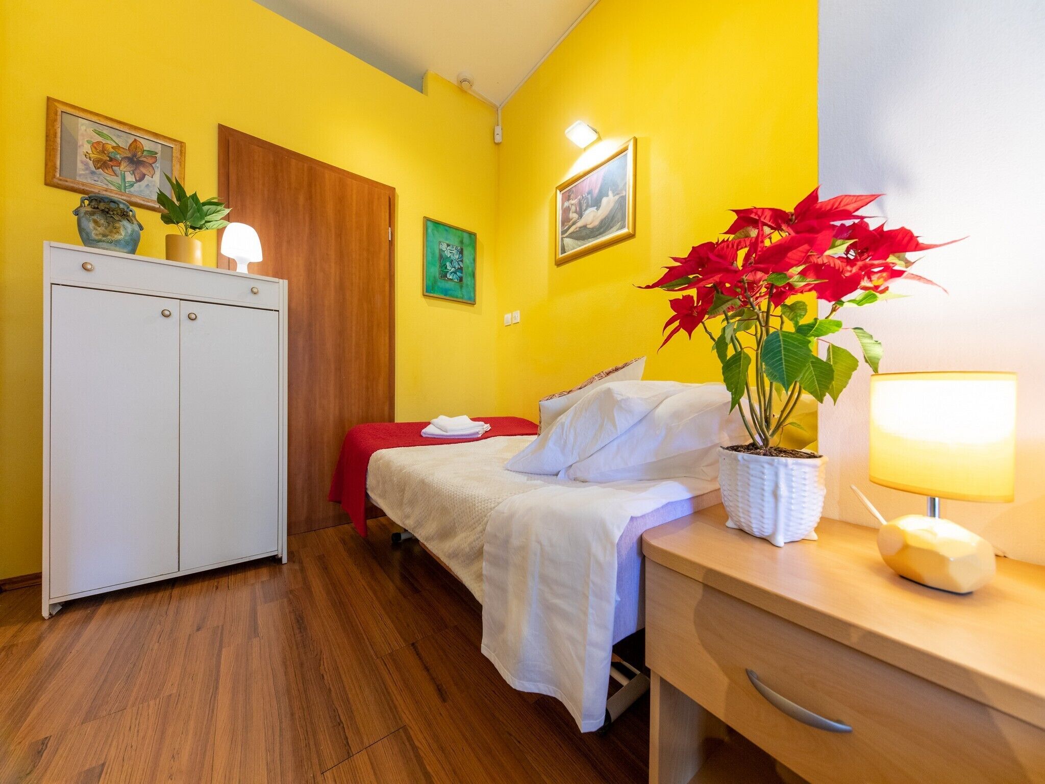 A hotel room with a wooden floor, a bed with white bedding, a desk with a lamp, and a potted plant with red flowers.