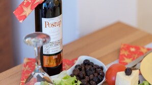 A bottle of Postup wine, a glass, a bowl of grapes, a tomato, a piece of cheese, and some meat on a wooden table.