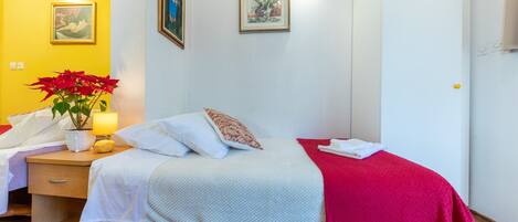 Quarto de hotel com cama, mesa de cabeceira, abajur, planta em vaso e duas imagens emolduradas na parede.