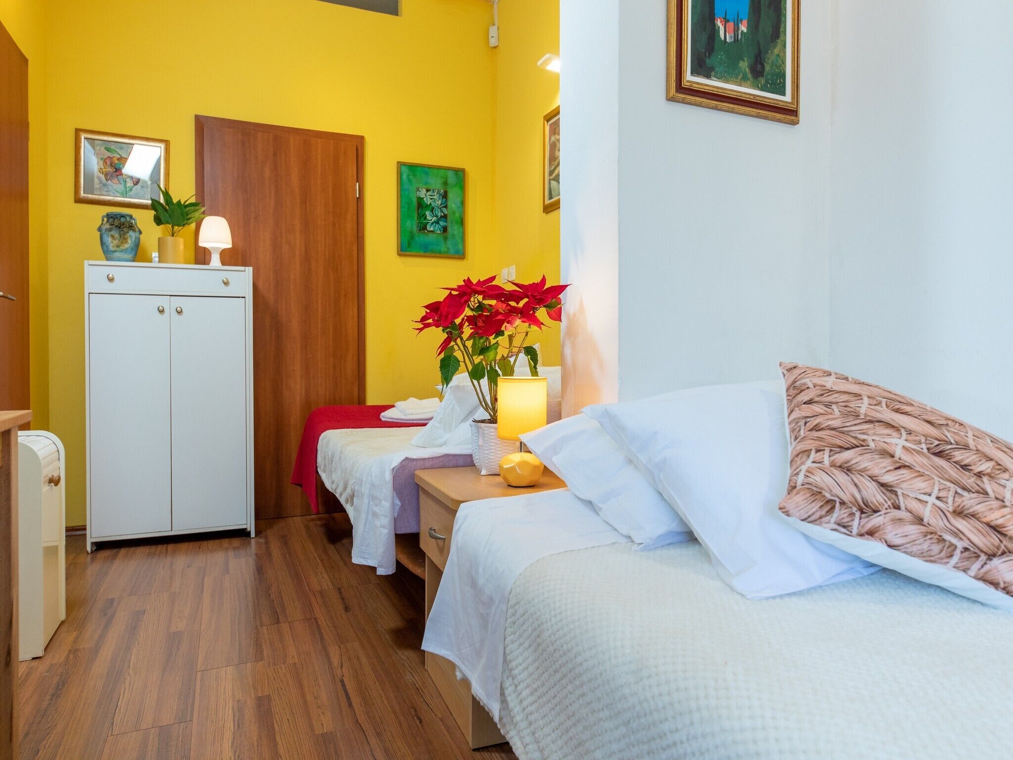 Quarto de hotel com guarda-roupa de madeira, cama com roupa de cama branca, mesa de cabeceira com abajur e uma cadeira vermelha.