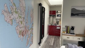 A compact hotel room with a kitchenette, a bed with a neatly folded towel, a map on the wall, and a painting above the bed. - Piper's Hut - Central apartment  (Portree)