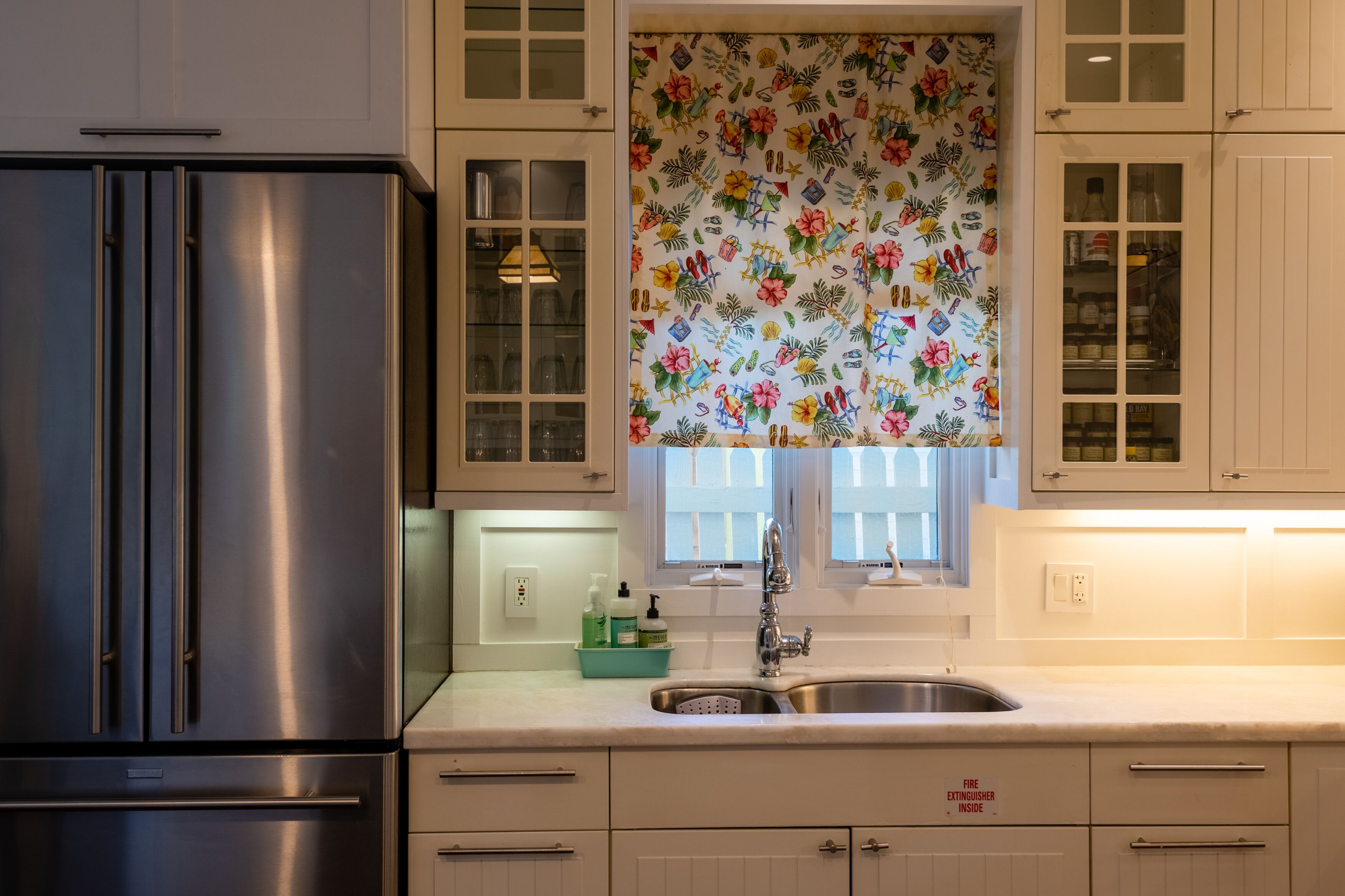 Een keuken met witte kastjes, een roestvrijstalen koelkast, een raam met een bloemen gordijn en een spoelbak met een bordje voor een brandblusser.