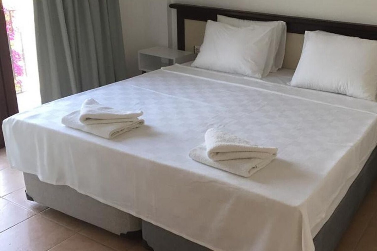 A neatly made bed with white linens and towels, a bedside table with a lamp, and a nightstand with a phone.