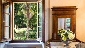 A room with a large window offering a view of a garden, a wooden cabinet with a mirror, and a vase with flowers on the counter. - Villa Turri is a historic mansion near Lucca in the heart of Garfagnana. (Filicaia)