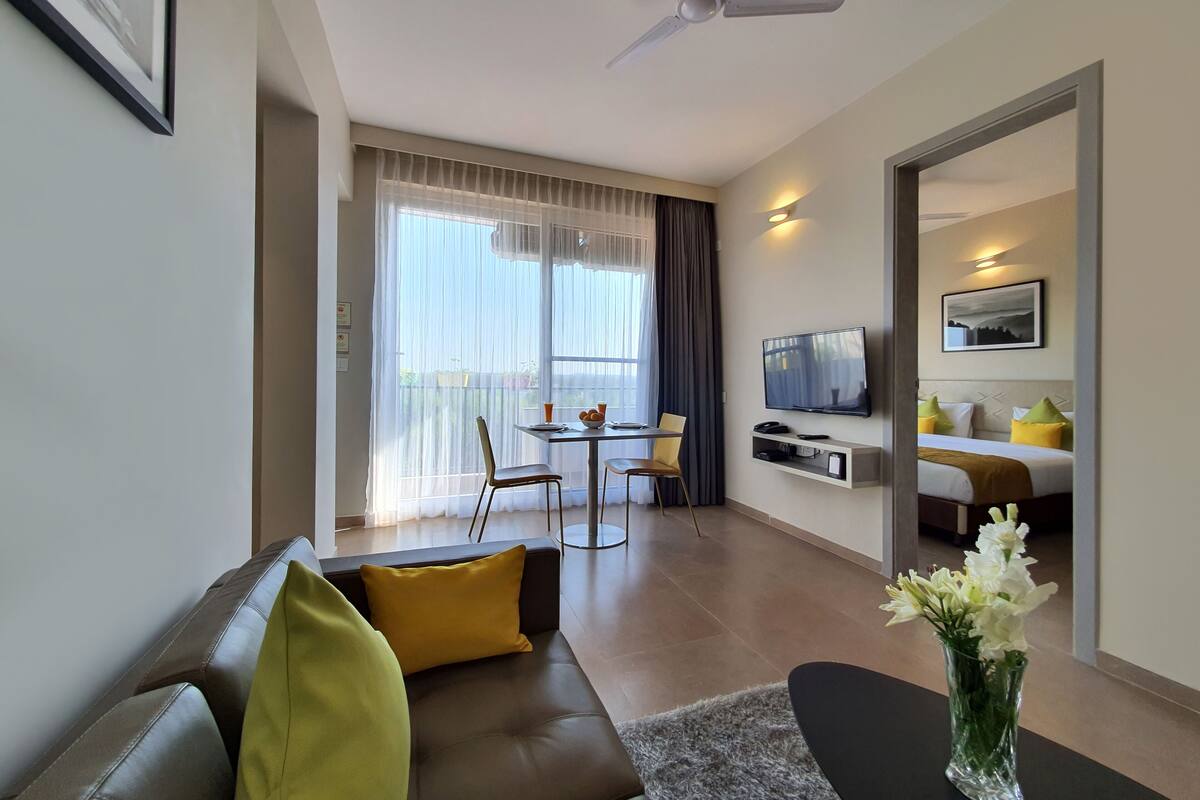 A modern hotel room with a sofa, a dining area with a table and chairs, a flat-screen TV mounted on the wall, and a bed with pillows.