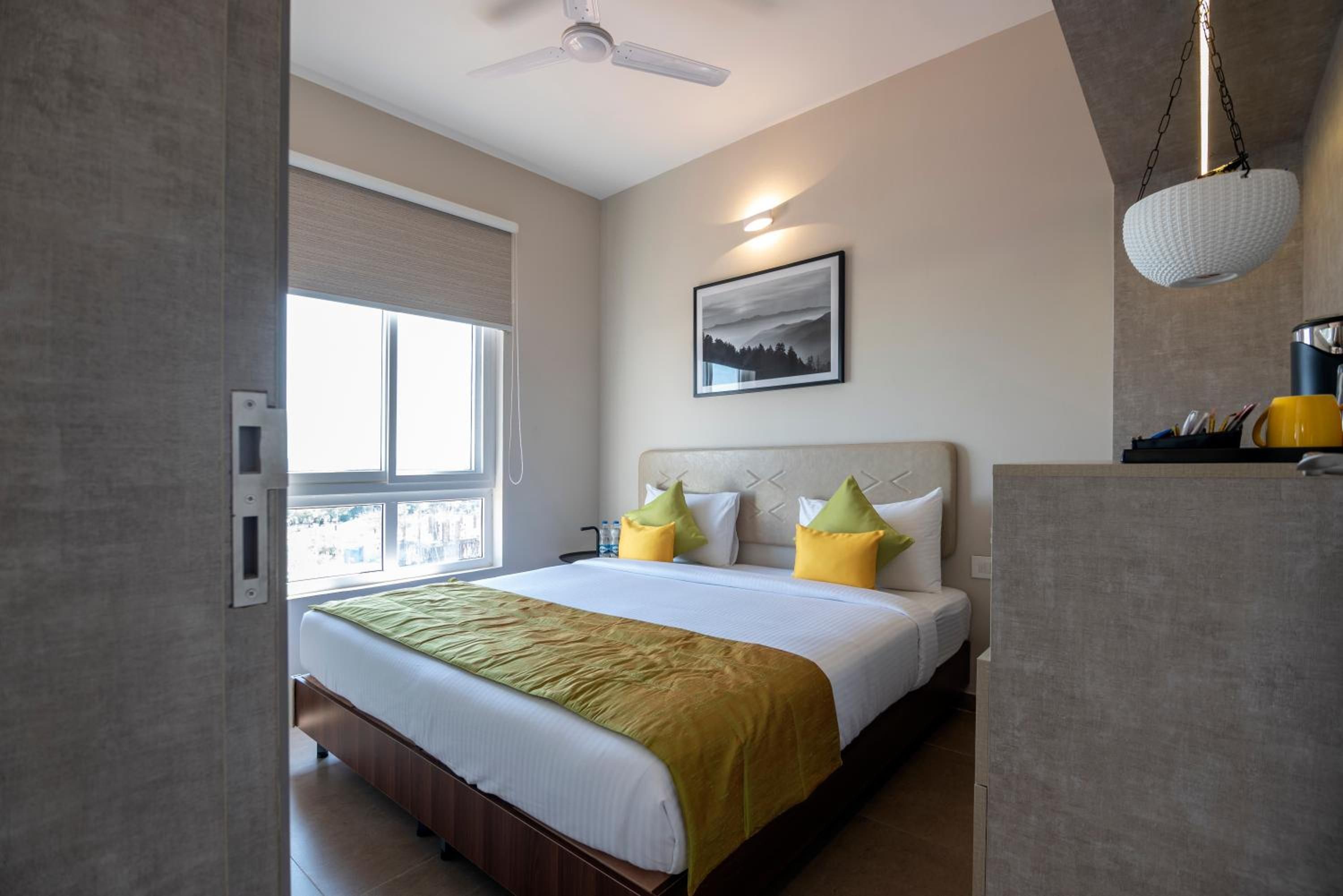 A hotel room with a bed, a window, a ceiling fan, and a painting on the wall.