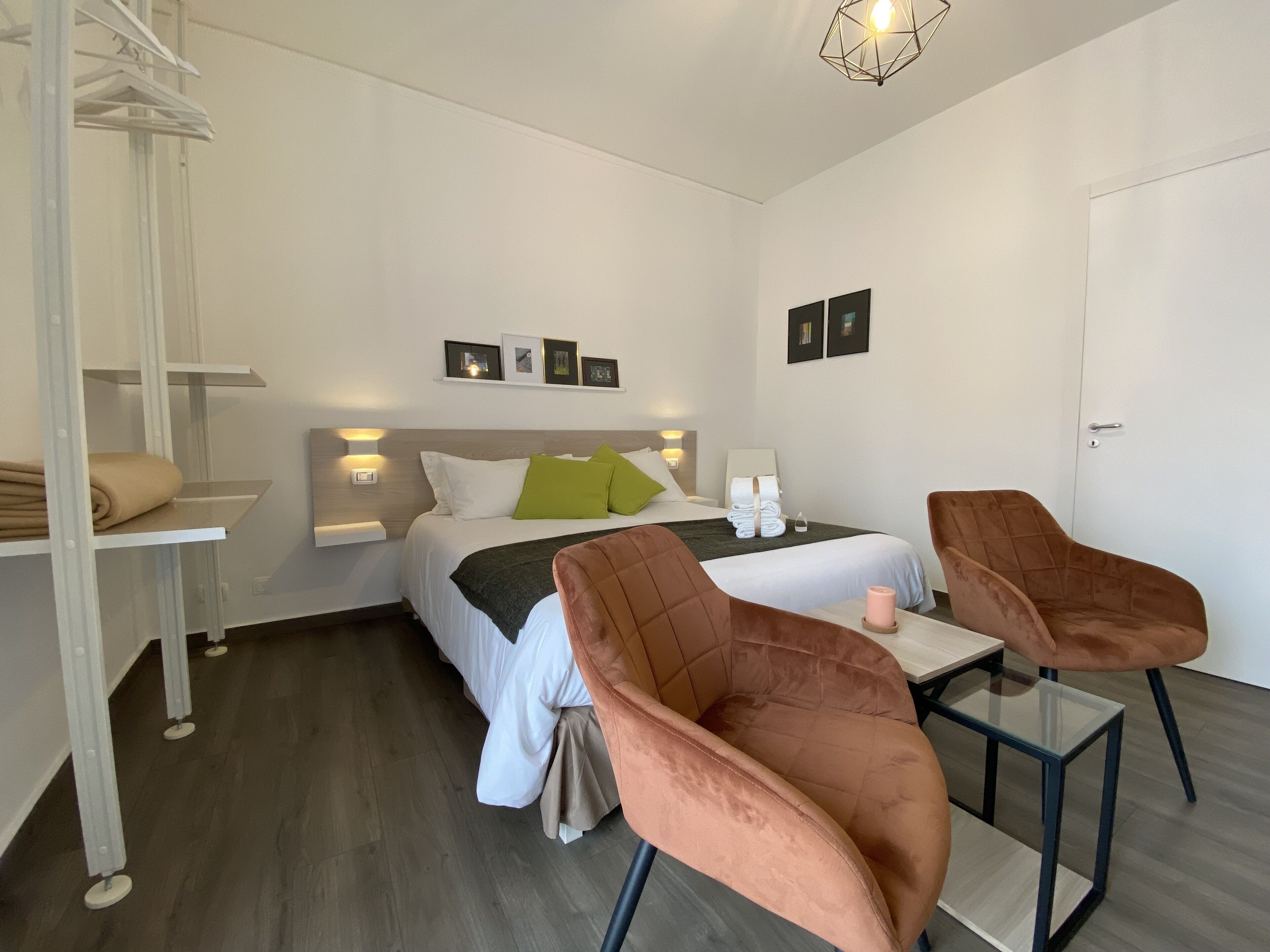 Ein modernes Hotelzimmer mit einem Bett, zwei Stühlen, einem kleinen Tisch, einer Wandablage mit Handtüchern und gerahmten Bildern an der Wand.