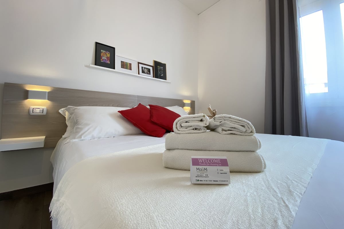 A hotel room with a neatly made bed, a welcome card, and a wall-mounted light fixture.