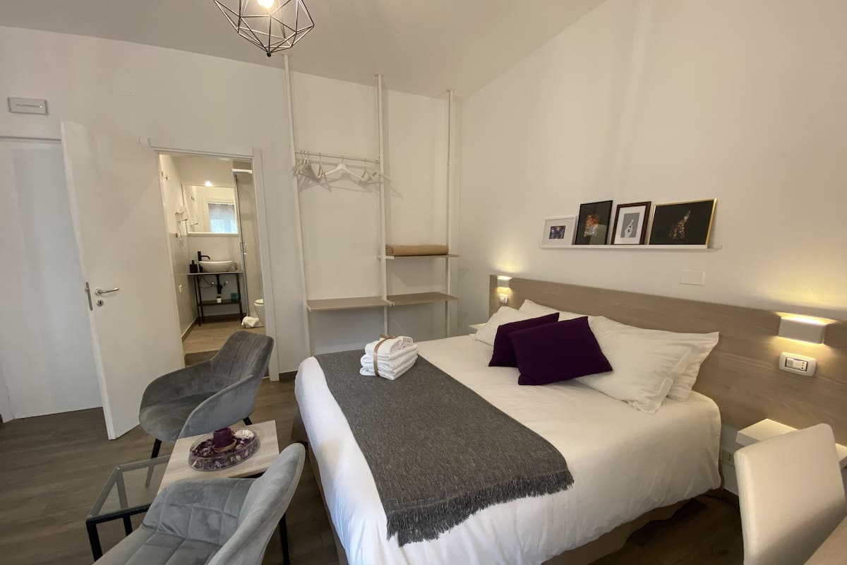 A modern hotel room with a bed, a chair, a small table with a decorative item, a bedside table, a wall-mounted shelf, and a bathroom visible through an open door.