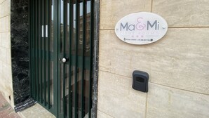 A building entrance with a green metal gate, a sign for 'Ma&Mi Light Rooms' with contact numbers, and a small black object on the wall. - Ma&Mi Light Rooms (Cefalù)