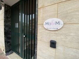 Entrada de un edificio con una verja metálica verde, un cartel que dice 'Ma&Mi Light Rooms' con números de contacto, y un objeto negro pequeño en la pared.