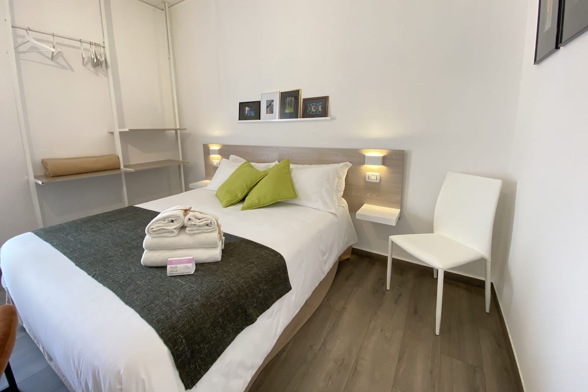 A neatly made bed with a rolled towel and a welcome sign, a white chair, and a wall-mounted shelf with hanging clothes.