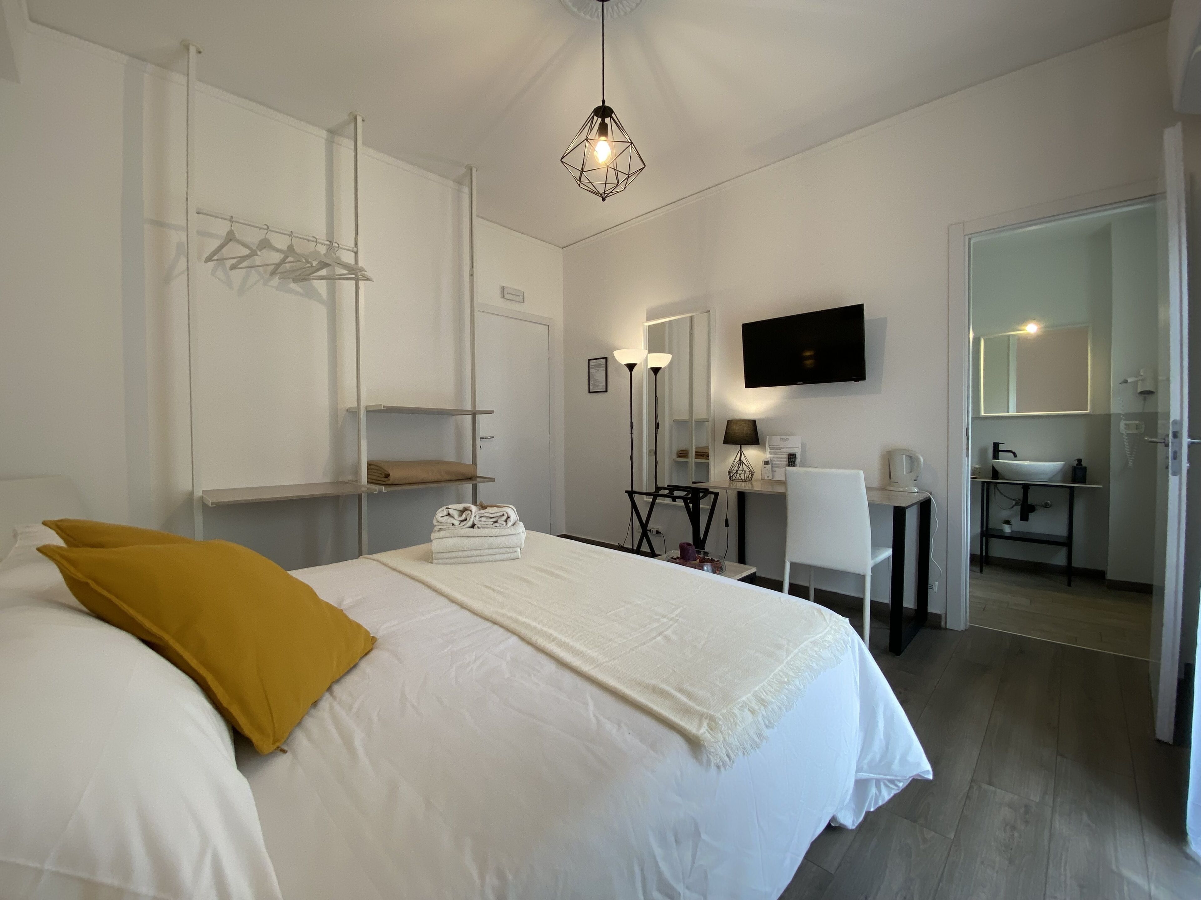 Habitación de hotel con cama, escritorio, silla, televisor, espejo y baño visible a través de una puerta abierta.