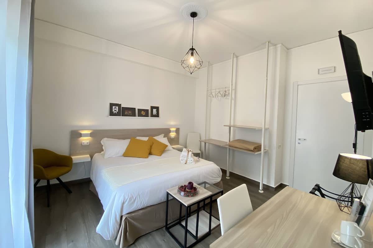 A hotel room with a bed, a desk, a chair, a hanging light fixture, and a coat rack.