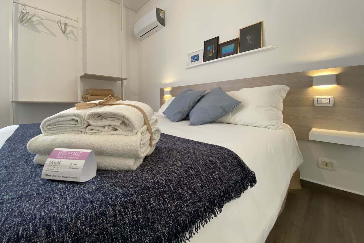 A neatly made bed with a folded towel and a welcome card, a bedside table with a lamp, and a wall-mounted air conditioner.