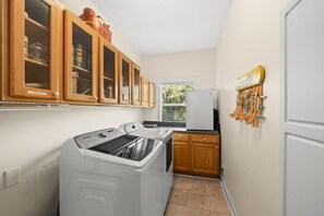 A compact kitchen with wooden cabinets, a washing machine, a refrigerator, and a window with blinds.