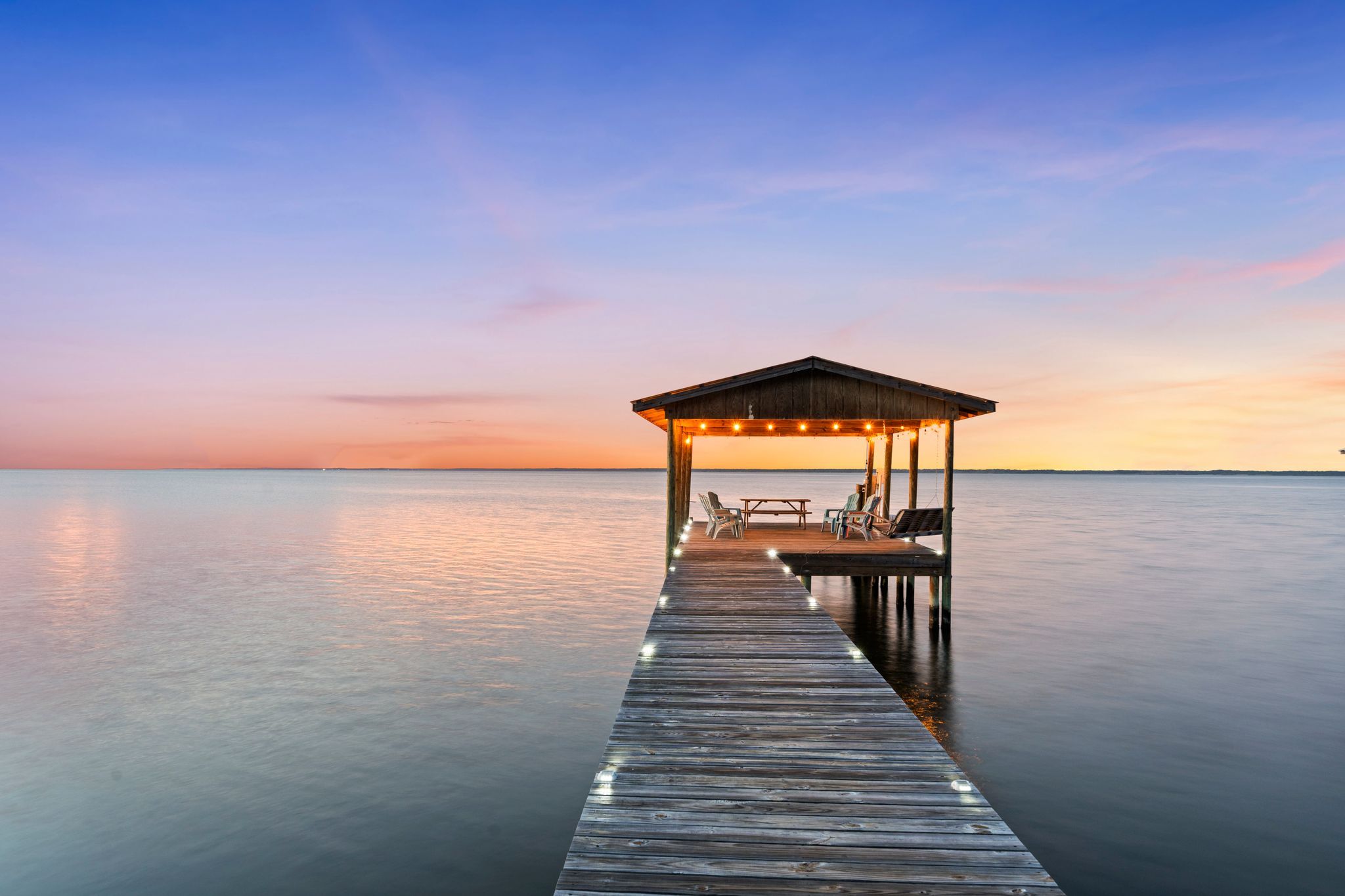 A wooden pier leading to a covered seating area with chairs and a table, overlooking calm water.