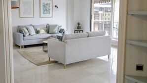 A modern living room with a grey sofa, a small side table, and a large window offering a view of buildings. - Superbe 120m2 - 2chambres + 2sdb, 3 Grds Balcons (Annecy)