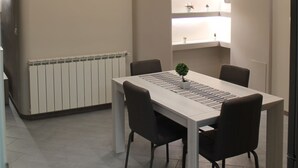 Un espace repas moderne avec une table blanche et quatre chaises, un luminaire au plafond et une étagère intégrée agrémentée d’objets décoratifs.
