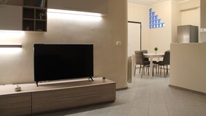 Un salon moderne avec un téléviseur à écran plat posé sur une console en bois, un coin repas avec une table et des chaises blanches, et un coin cuisine équipé d’un réfrigérateur et de placards.