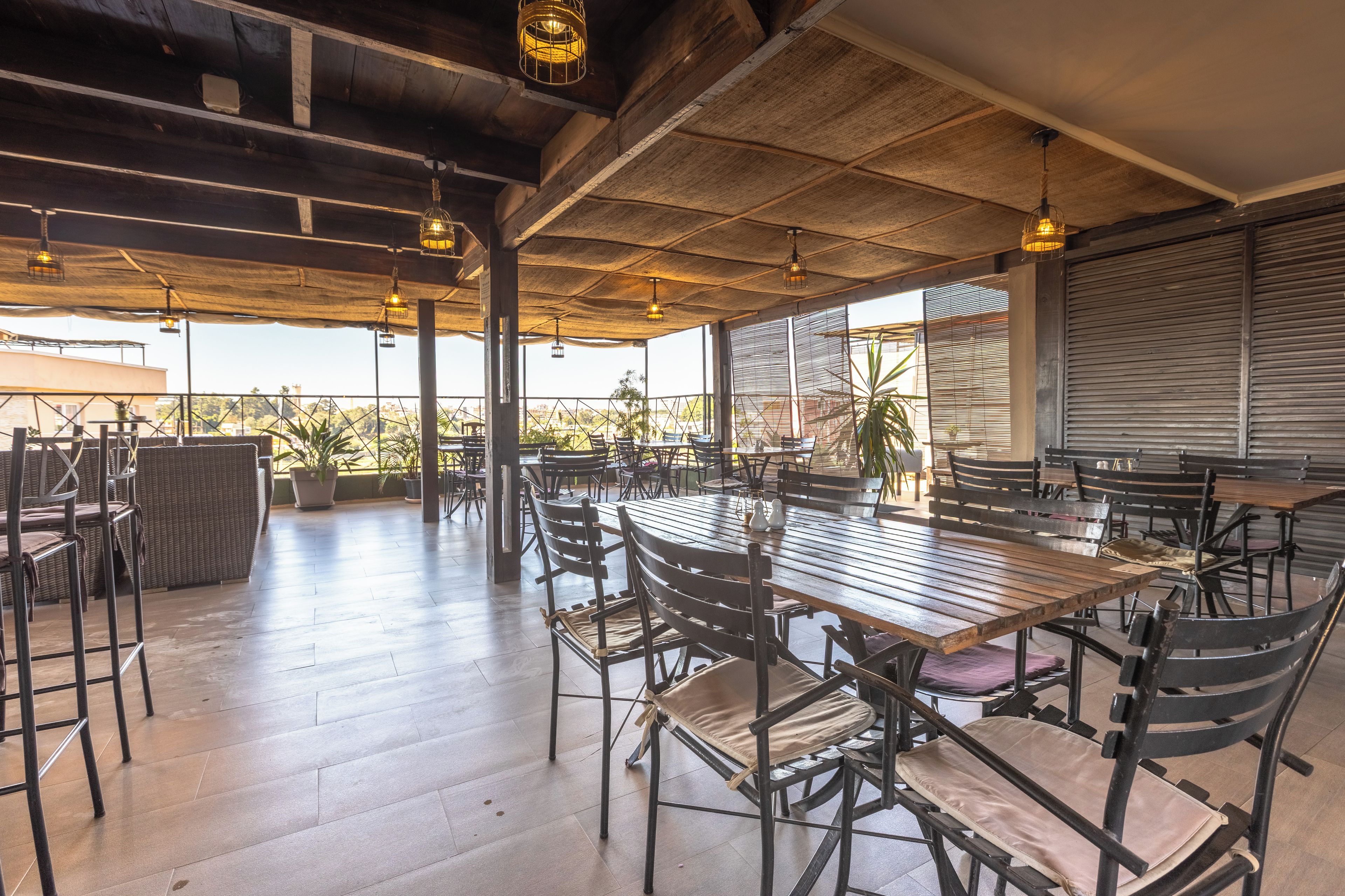 An outdoor dining area with wooden tables and chairs, potted plants, and a view of the cityscape.