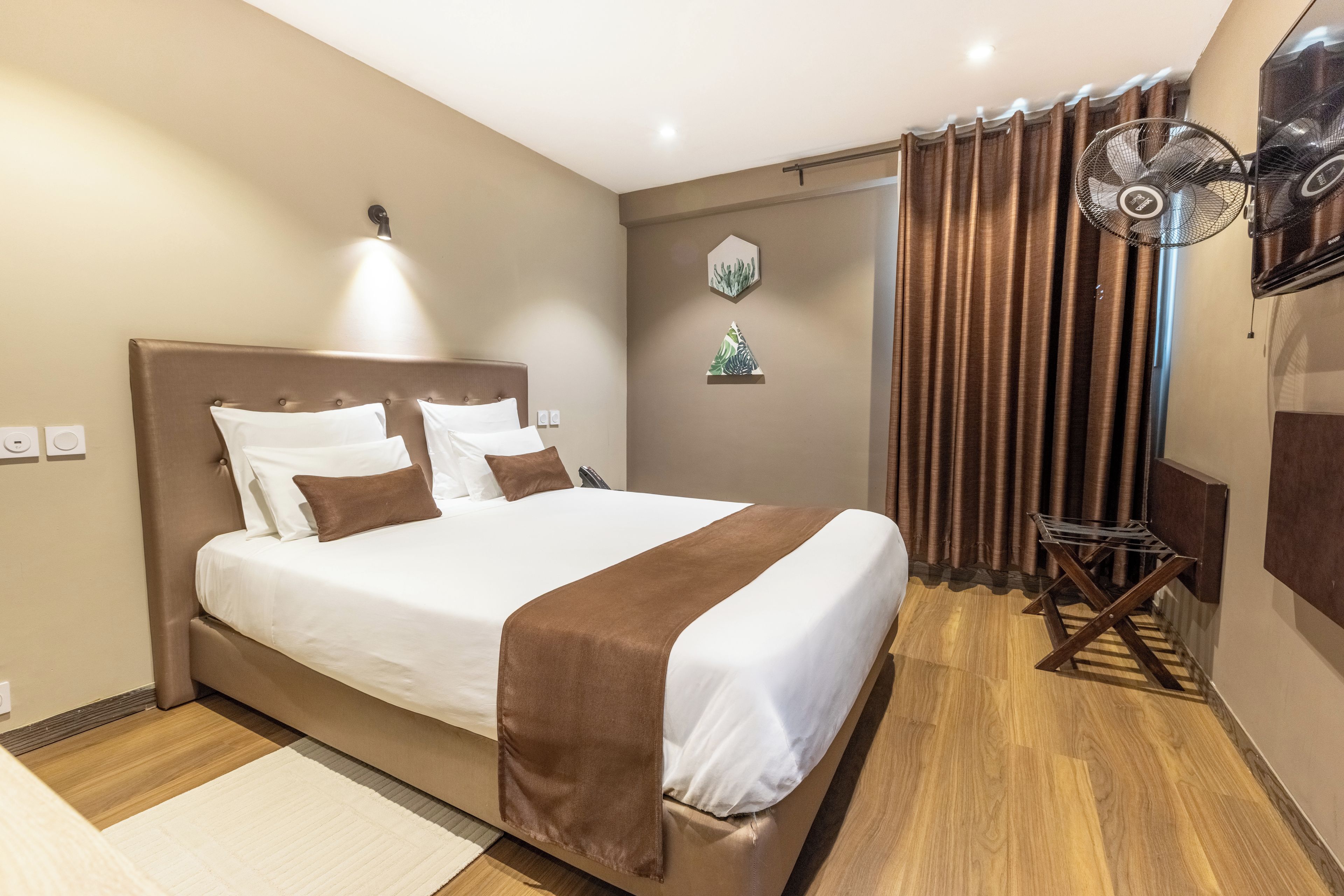 A hotel room with a large bed, a bedside table, a wall-mounted fan, and a window with curtains.