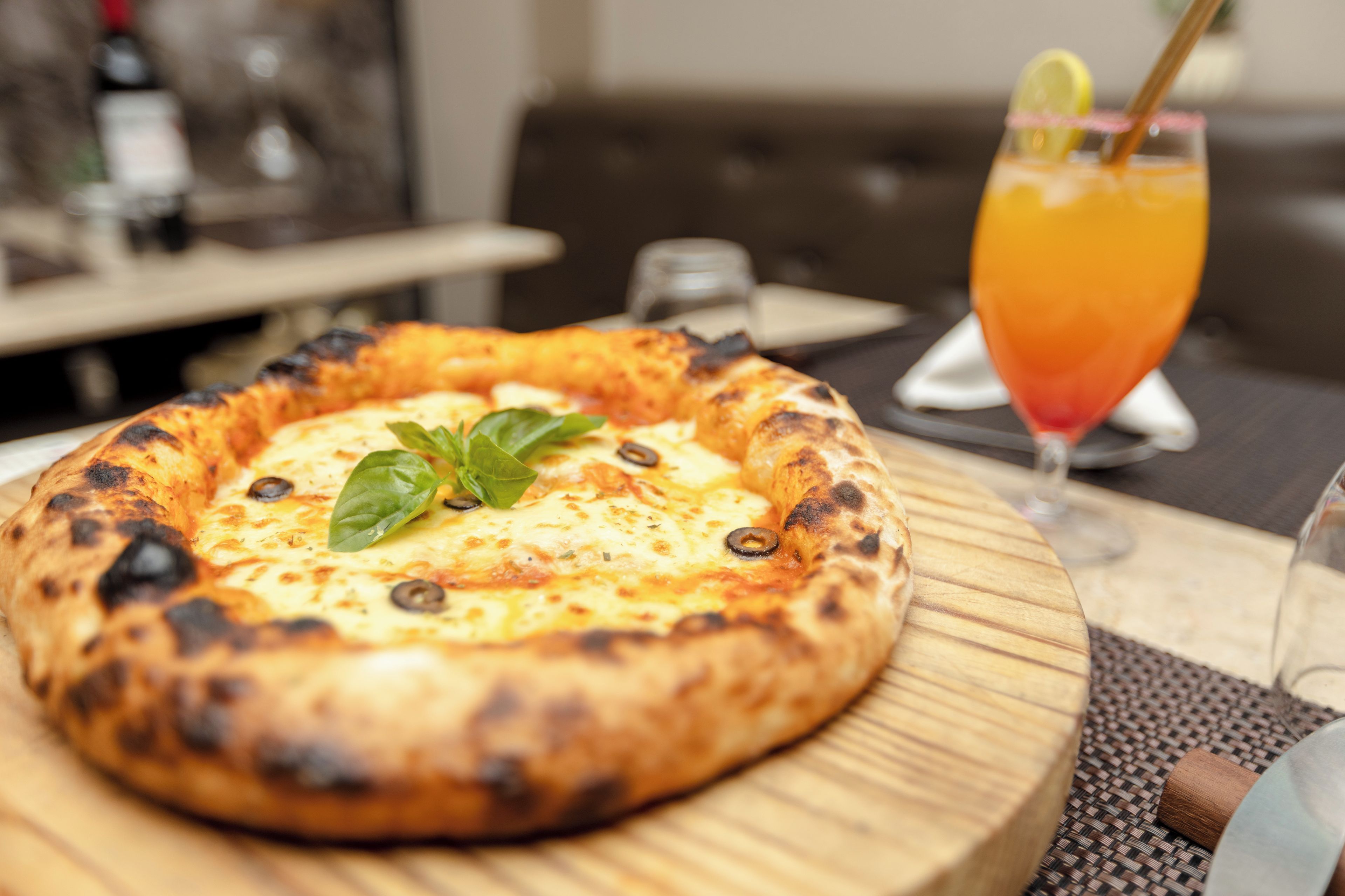 A pizza with a crispy crust, topped with cheese, olives, and a basil leaf, served on a wooden board.