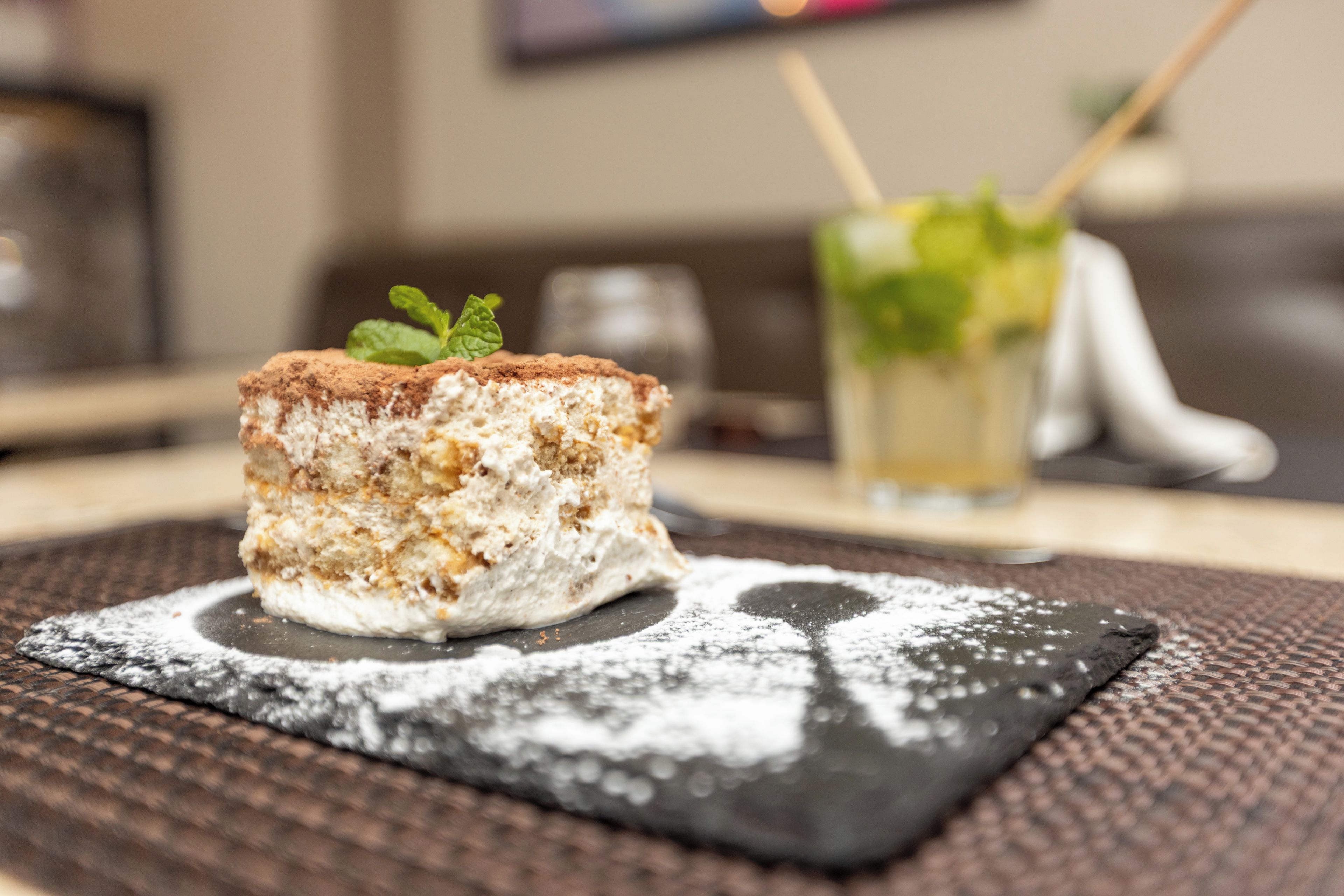 Un dessert servi sur une assiette carrée, saupoudré de sucre glace, accompagné d’un verre décoré d’une feuille de menthe, le tout sur un fond flou.