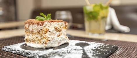 Postre sobre un plato cuadrado con azúcar glasé, un vaso con adorno de menta y un fondo desenfocado.