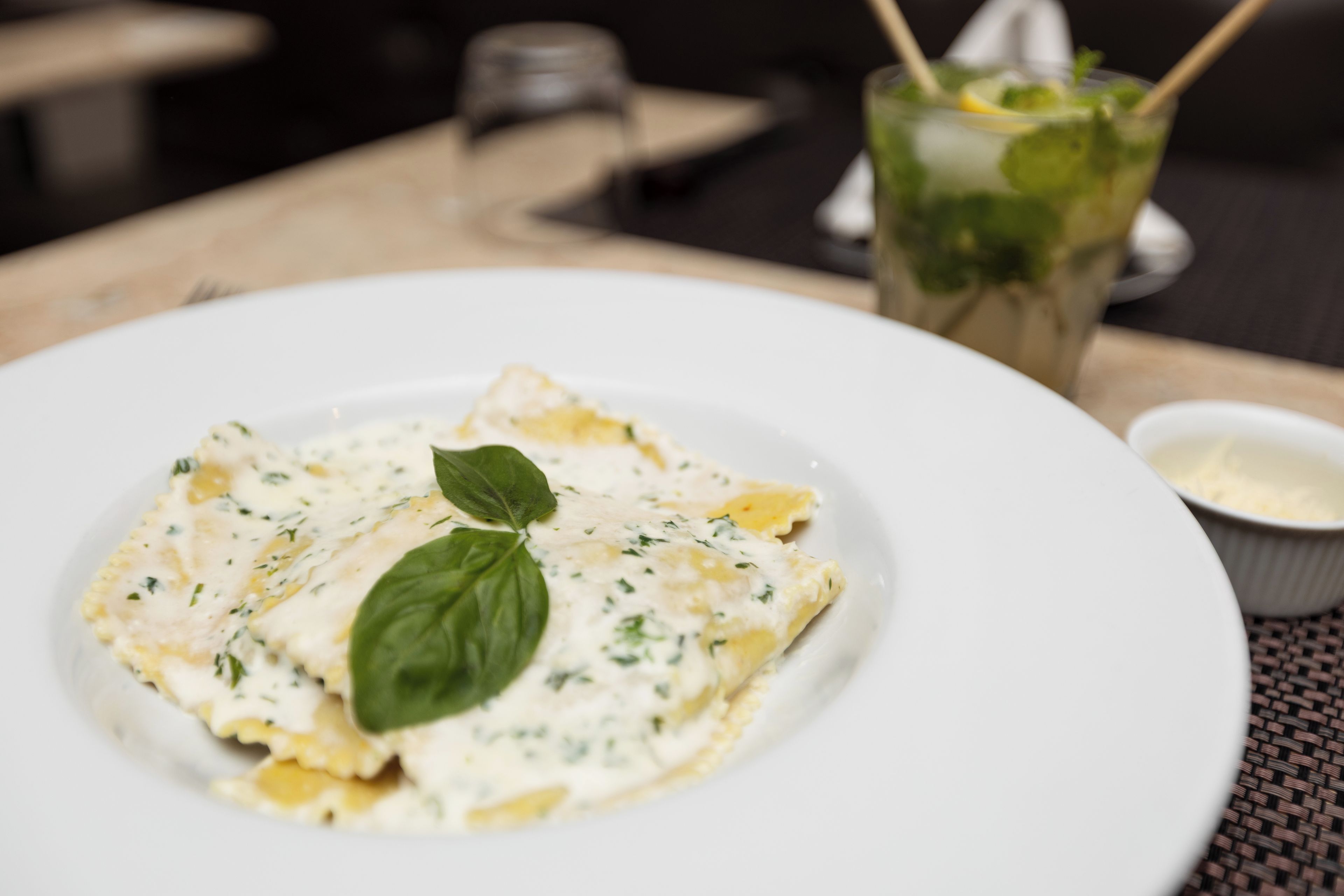 A plate of food with a basil leaf garnish, a glass of drink with a straw, and a small bowl of sauce on a table.