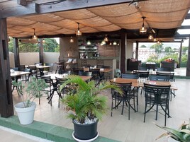 Un área de café al aire libre con mesas y sillas de madera, plantas en macetas e iluminación colgante.