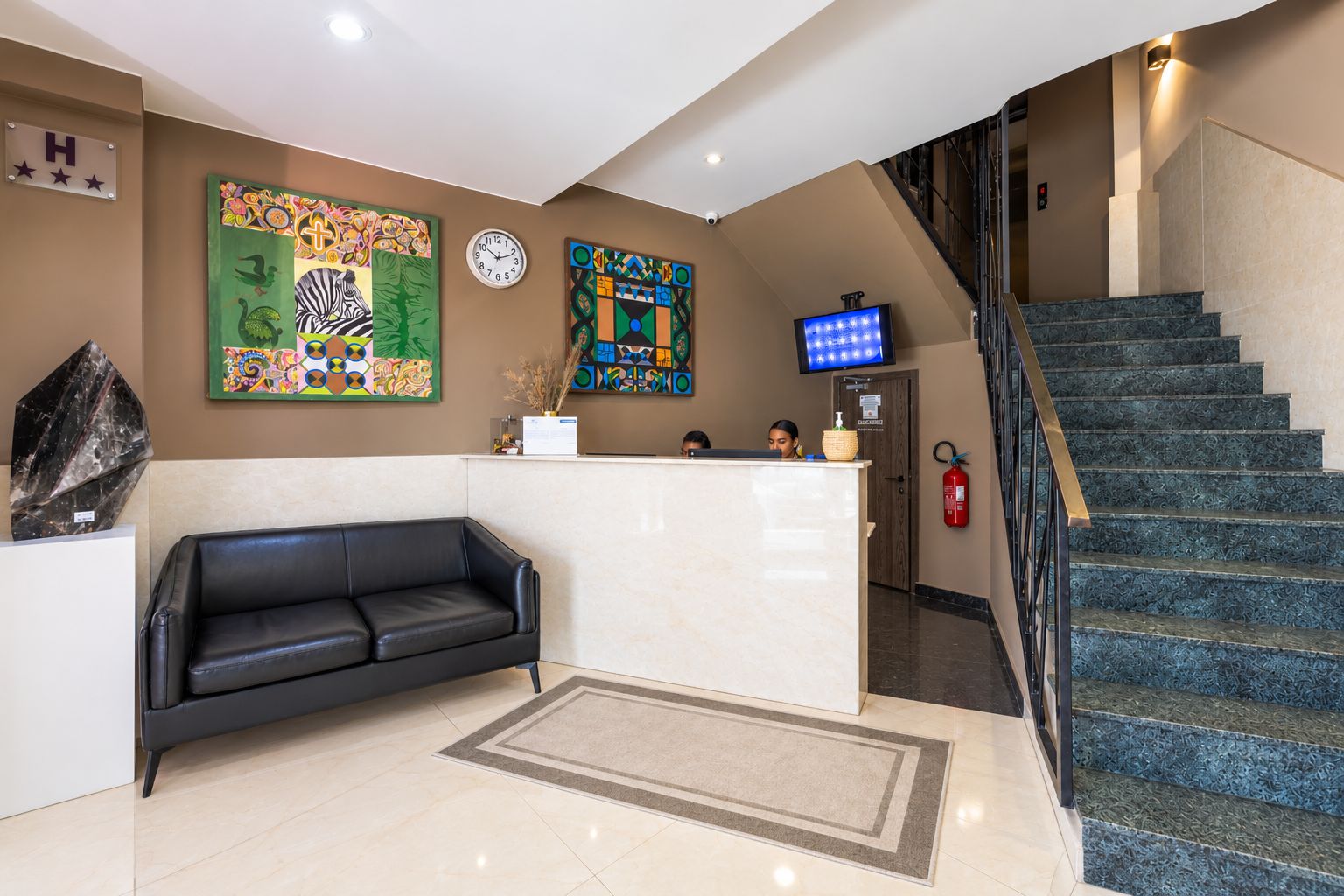 Un hall d’hôtel comprenant un comptoir d’accueil, un canapé en cuir noir, une horloge et un escalier.