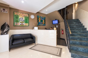 A hotel lobby with a reception desk, a black leather couch, a clock, and a staircase. - Fly Inn Madagascar Hotel (Antananarivo)