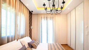 A modern bedroom with a large bed, a chandelier, and a view of the outdoors through the windows. - Perla Nera | Heated Pool (Georgioupoli)