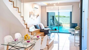 A modern living room with a glass dining table, white sofas, and a view of a swimming pool. - Perla Nera | Heated Pool (Georgioupoli)