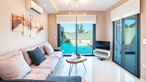 A modern living room with a sofa, TV, and a pool view. - Perla Nera | Heated Pool (Georgioupoli)