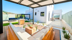 A modern outdoor patio with a built-in grill, seating area, and a view of the surrounding landscape. - Perla Nera | Heated Pool (Georgioupoli)
