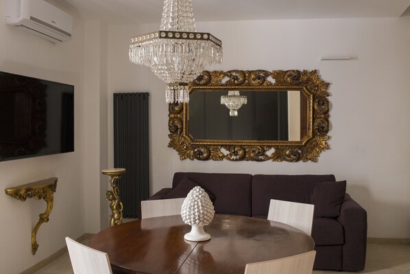 Un coin repas avec une table ronde, un objet décoratif blanc, un canapé marron foncé, un miroir avec un cadre doré et un lustre.