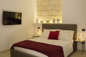 A neatly made bed with a red throw, a flat-screen TV on the wall, and a brick-patterned wall behind the bed. - Le Dimore del Centro (Noto)