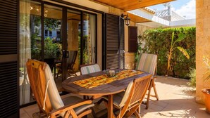 Een terras buiten met een houten tafel en stoelen, omringd door glazen deuren en uitzicht op groen.