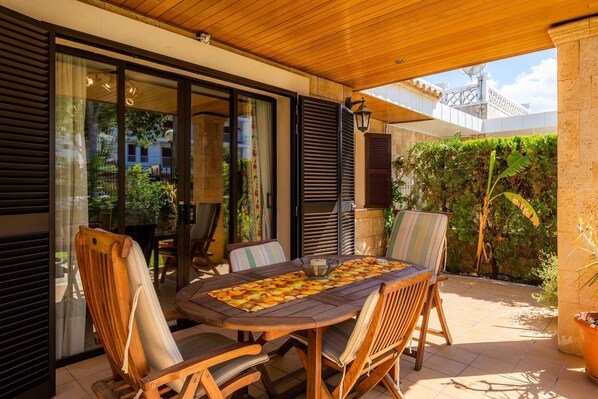 An outdoor patio with a wooden table and chairs, surrounded by glass doors and a view of greenery.