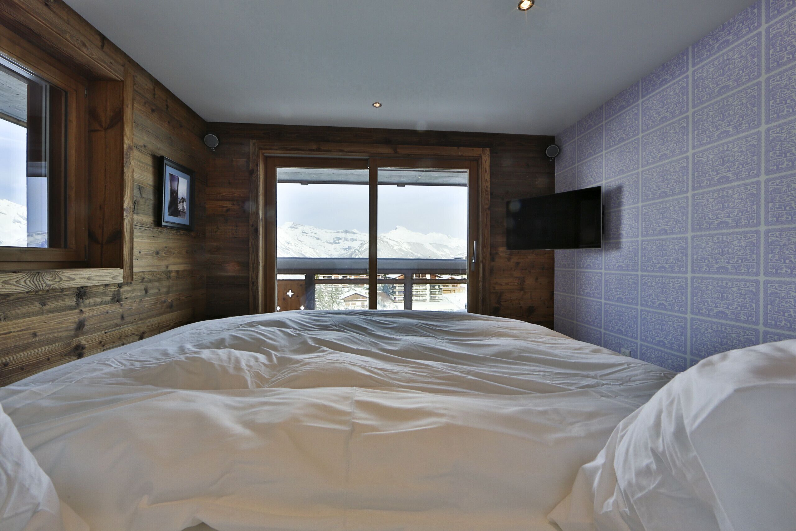 A bedroom with a large bed, wooden walls, a window with a mountain view, and a mounted TV.