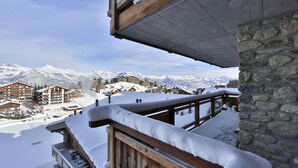 Un balcone in legno con ringhiere imbiancate dalla neve che si affaccia su una località turistica montuosa con diversi edifici.