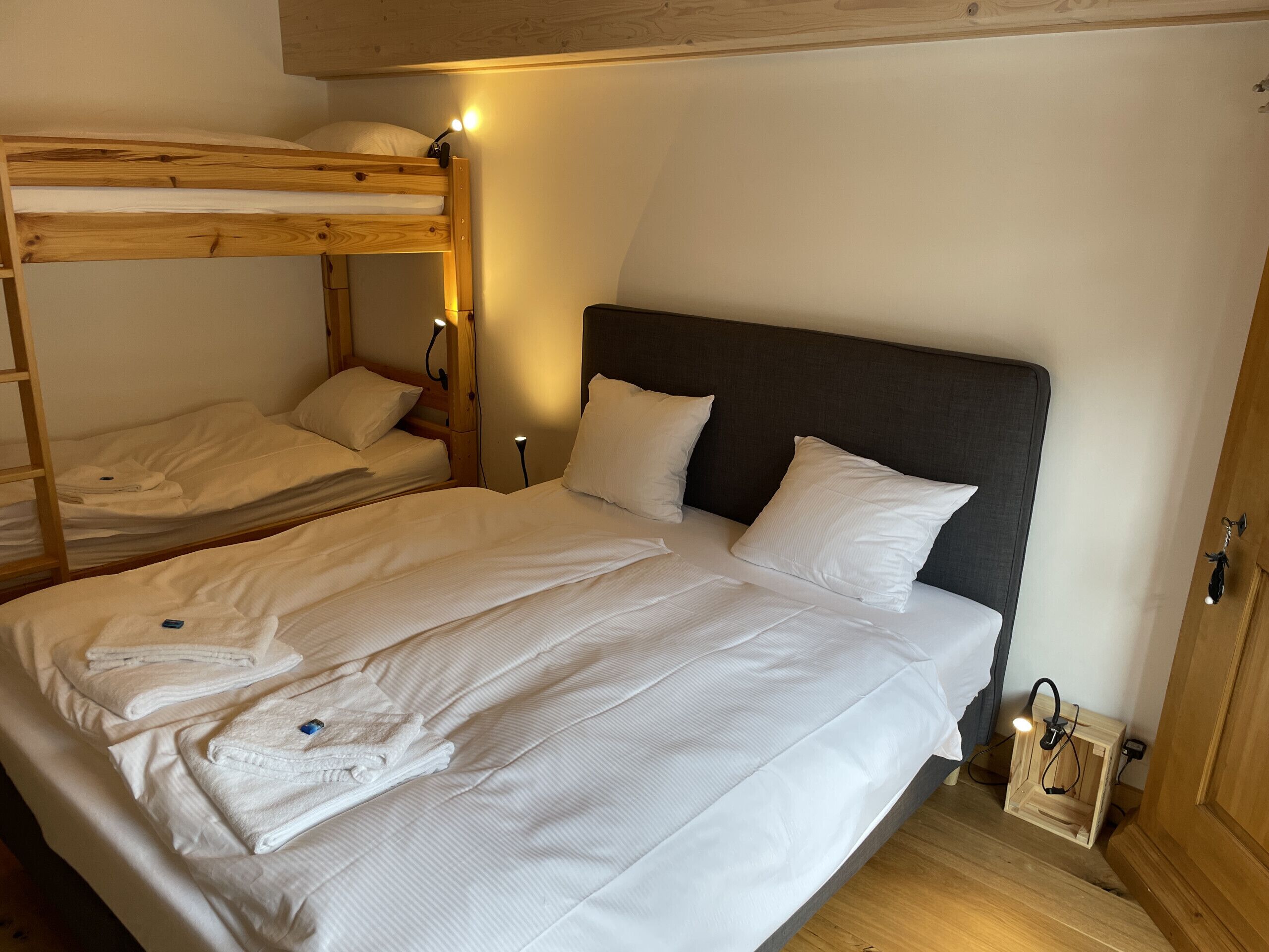 Een kamer met een stapelbed en een enkel bed, beide met witte lakens. Er staan nachtlampjes en een klein houten nachtkastje met een lamp.