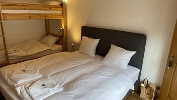 Een kamer met een stapelbed en een enkel bed, beide met witte lakens. Er staan nachtlampjes en een klein houten nachtkastje met een lamp.