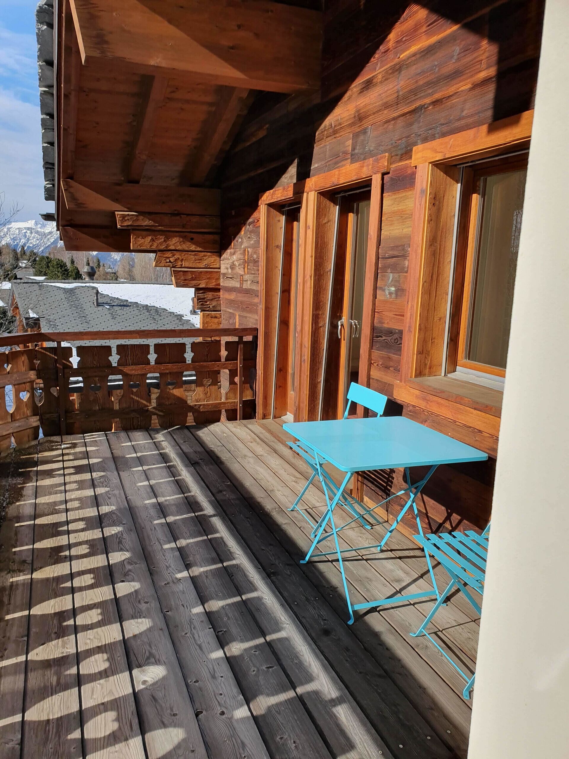 Eine Holzterrasse mit blauem Tisch und Stühlen, ein Holzzaun und im Hintergrund eine Aussicht auf schneebedeckte Berge.