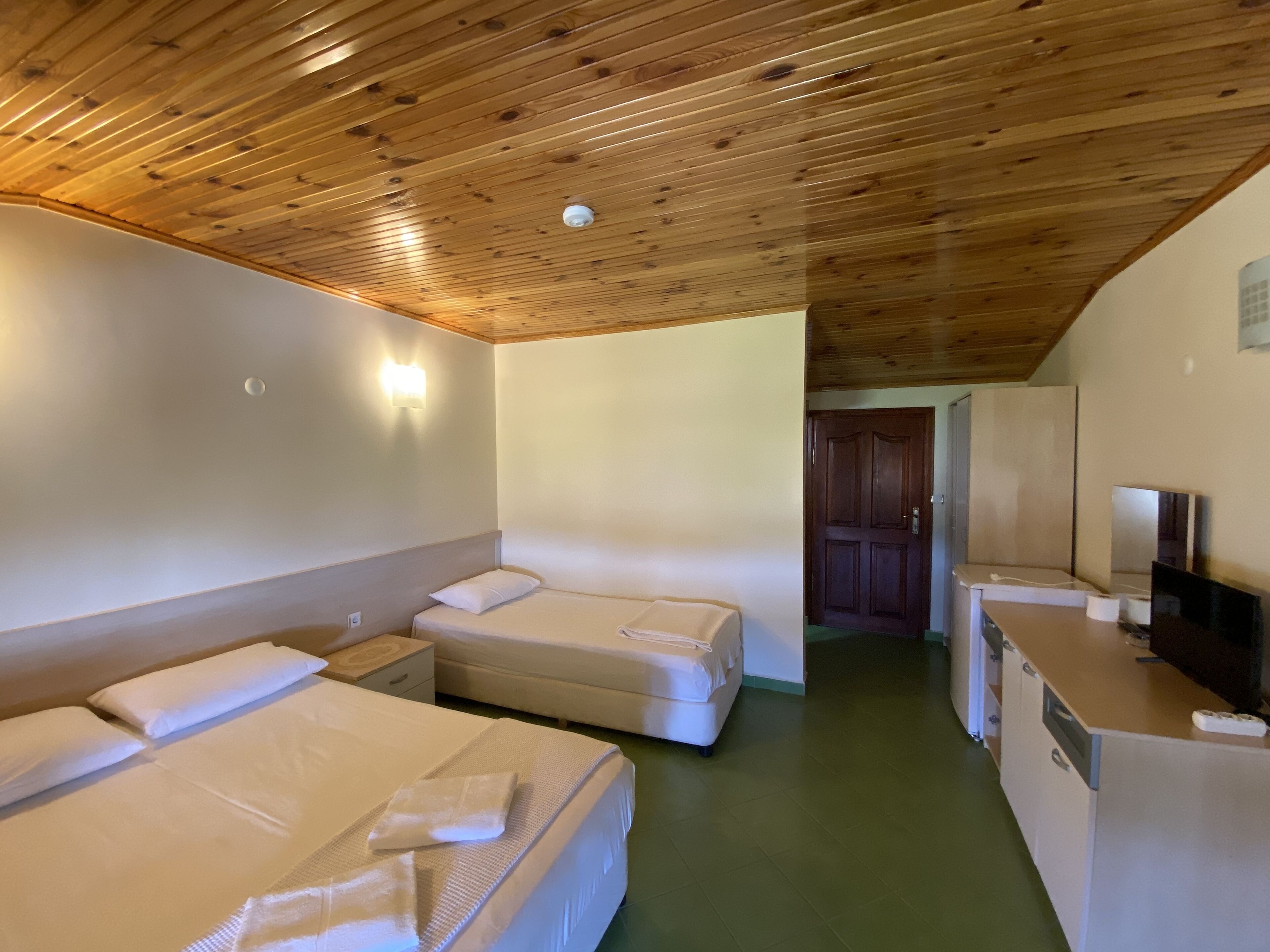A room with two beds, a wooden ceiling, a small desk with a TV, and a door leading to another room.