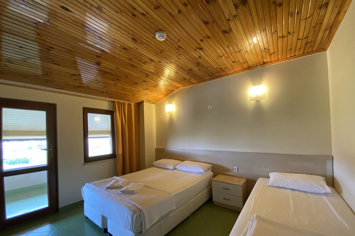 A room with two beds, a window with blinds, a wooden ceiling, and a wall-mounted light fixture.
