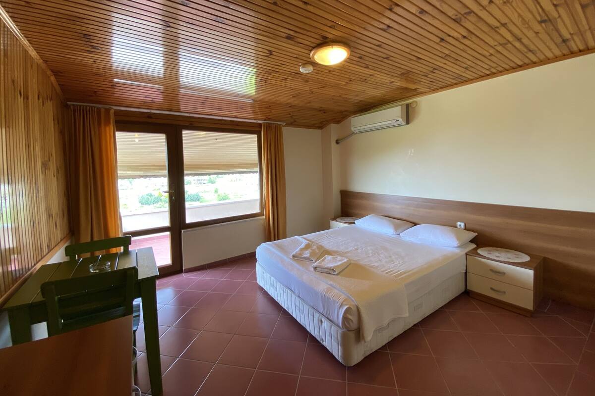 A room with a wooden ceiling, a bed with white linens, a nightstand, and a window with curtains.